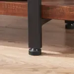 Close-up of a black metal furniture leg with an adjustable leveling foot from the 05.03 Solid Wood Bookcase, a 5-tier industrial rustic vintage shelf in distressed brown, resting on a wooden floor by the bookcase.