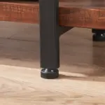 Close-up of a black metal furniture leg with adjustable leveling foot on light wood flooring, next to the 05.03 Solid Wood Bookshelf, a 3-tier rustic vintage industrial etagere bookcase in distressed brown finish.