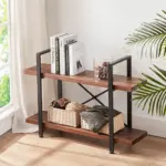 The 07.03 Solid Wood Bookcase, a 2-tier industrial rustic vintage etagere bookshelf in distressed brown, stands by a window and potted plant on a light wood floor, holding books, a glass candle holder, baskets, and folded blankets.