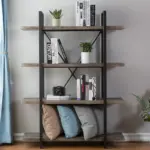 The 4 Tier Bookcase, displayed against a light-colored wall near a window, is styled with books, potted plants, a camera on the shelves, and three cushions on its bottom shelf.