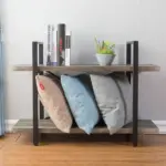 The BC02-01 07 - 03 2-Shelf Rustic Industrial Bookshelf, made of wood and metal, displays books and a potted plant on top, while three pillows rest on the lower shelf against a light gray wall—ideal for ODM/OEM projects.