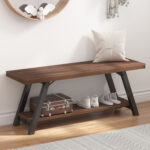 The Industrial Wood Bench, Walnut features black metal legs and a walnut finish, shown on a hardwood floor with a white pillow on top and shoes, a suitcase, and a book neatly placed on the lower shelf.