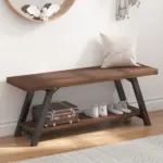 The Industrial Wood Bench, Walnut features black metal legs and a wooden seat with a white pillow. Its lower shelf displays two pairs of shoes and a brown suitcase, blending style with practical walnut furniture accents.