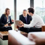 Three business professionals sit on couches in an office lounge, discussing information about the OEM Service Process displayed on a digital tablet.