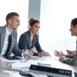 Four people in business attire sit around a conference table, engaged in discussion with documents and electronic devices in front of them.