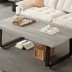 The Minimalist Coffee Table, Light Grey Oak, 47 Inch features black legs and displays a tray with a teapot and cups, a sculpture, and books on top; underneath are a wicker basket and additional books.