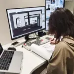 Person working at a desk with two monitors, one displaying architectural designs, while writing notes on paper beside a laptop.