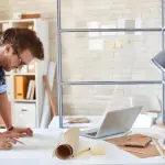 A man in a plaid shirt stands at a desk, working on architectural plans and reviewing the OEM Service Process next to a laptop, lamp, and office supplies in a modern workspace.