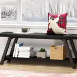 An Industrial Wood Bench in Black Oak sits in front of a snowy window, adorned with a red plaid pillow featuring a white reindeer, plus a cup and saucer, books, a plant, and a wicker basket.