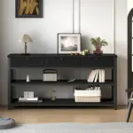 A Sofa Table with 3 Drawers, Black Oak, holds books, framed art, plants, a lamp, and decor items. A robot vacuum is nearby on the floor.