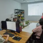 A group of people in an office meeting room watch a presentation with a spreadsheet projected on the wall. A presenter stands at the front, while others sit at a table with computers.