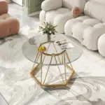 A glass-top coffee table with a gold geometric base holds a vase of flowers, a fruit bowl, and a magazine in a modern, light-colored living room.