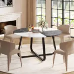A Round Dining Table for 4 to 6 People, featuring a 42.13" white and black faux marble top, is paired with four beige chairs and decorated with flowers, bottles, and a digital clock in a bright room with large windows.