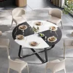 A 42.13" round dining table for 4–6 people with a white and black faux marble top is set with beige chairs, plates, glassware, cutlery, and a fruit bowl centerpiece.