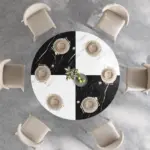 A 42.13" round dining table for 4 to 6, featuring white and black faux marble, is set with beige chairs, neutral dishware, and a small vase of green apples in the center.